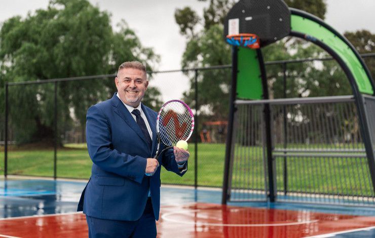 Smiling man posing on a sports court while holding a tennis racquet and tennis call.