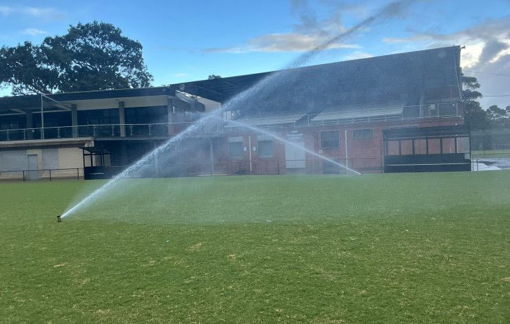 Two sprinklers spray water across a grass oval in front of a sports pavilion.