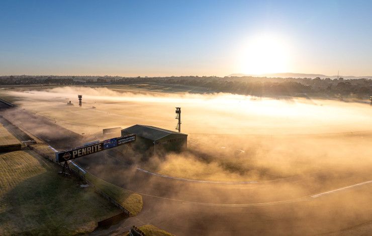 Looking down over the home straight of a racing track that is covered in mist as the sun rises.