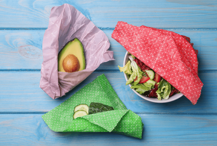 An avocado, a cucumber and a bowl of salad in colourful beeswax wraps on a blue background.