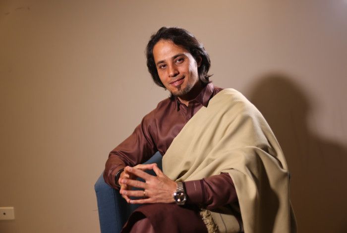 A man in a brown shirt, sitting with his fingertips resting together, looking peaceful, with a cream wrap over his shoulder