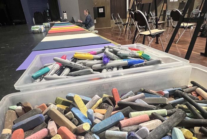 containers of coloured chalk sit in the forfront creating a persepctive view of a long table with legs of easels in the background. 