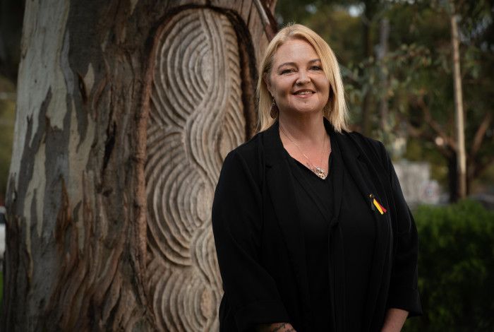 A smiling woman dressed in black stands in front of an Aboriginal message tree.