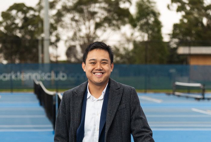 A smiling man leans on a tennis net.