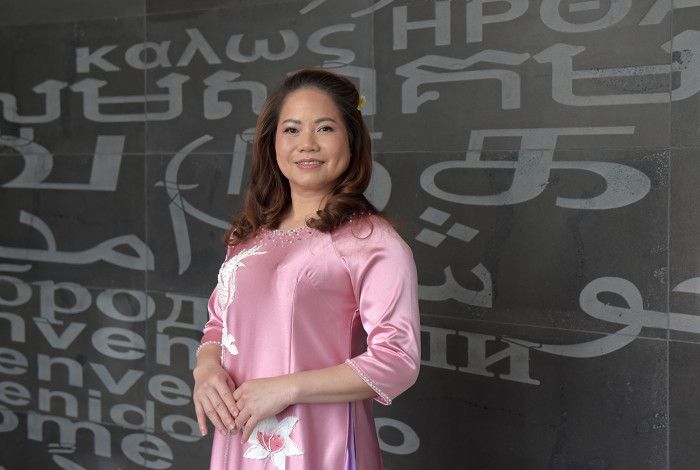 A smiling woman dressed in a pink silk dress in front of a black wall covered in grey lettering
