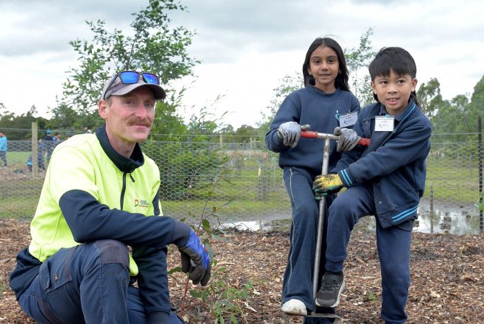 A man crouches next to two children who are helping him plant a tree