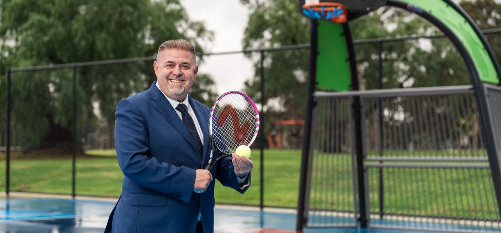 Smiling man posing on a sports court while holding a tennis racquet and tennis call.
