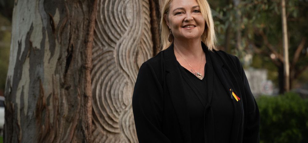 A smiling woman dressed in black stands in front of an Aboriginal message tree.