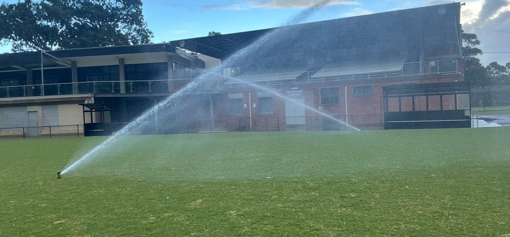 Two sprinklers spray water across a grass oval in front of a sports pavilion.
