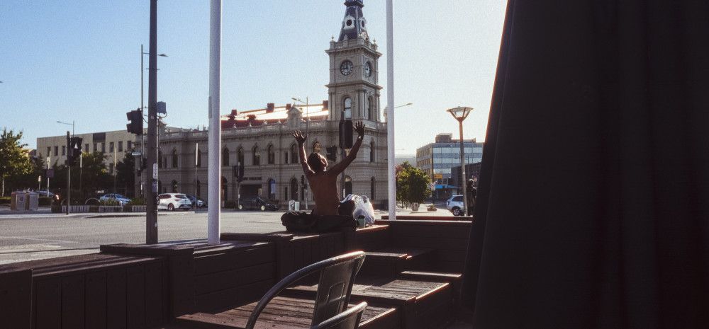 A man sits in the sunshine with his hands raised to the air across the street from a historical building and clock tower