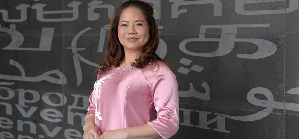 A smiling woman dressed in a pink silk dress in front of a black wall covered in grey lettering