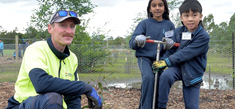 A man crouches next to two children who are helping him plant a tree