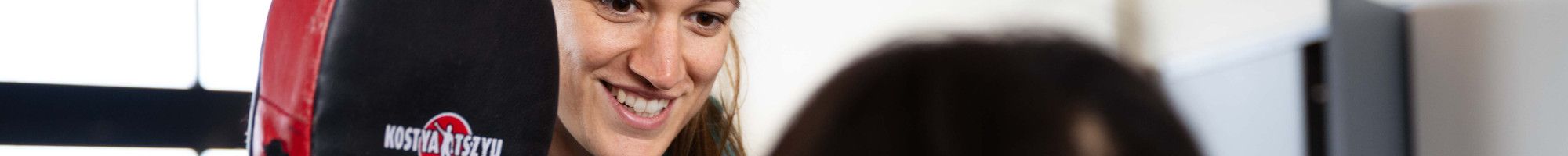 A smiling woman holds a boxing pad in front of someone standing with their back to the camera