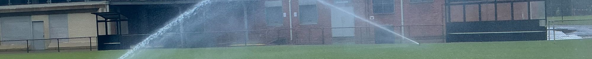 Two sprinklers spray water across a grass oval in front of a sports pavilion.