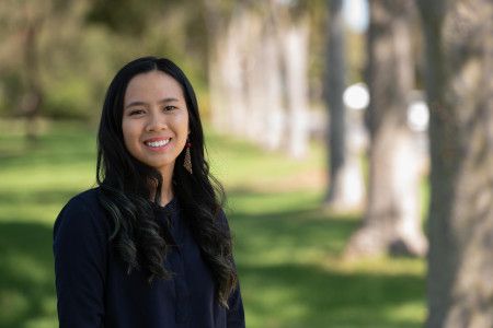 A woman with long dark hair smiles at the camera while standing in a park location
