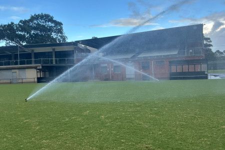 Two sprinklers spray water across a grass oval in front of a sports pavilion.