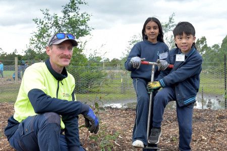 A man crouches next to two children who are helping him plant a tree