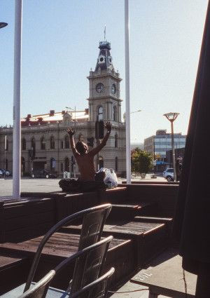 A man sits in the sunshine with his hands raised to the air across the street from a historical building and clock tower